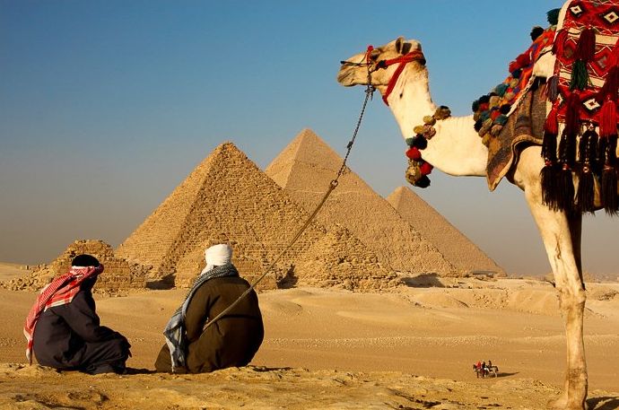 Dos beduinos descansando en la arena del desierto mirando las pirámides de Egipto mientras su camello espera